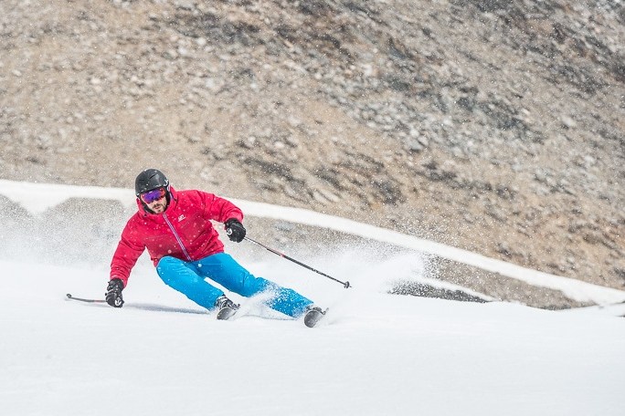 Skier in red jacket and blue pants skiing downhill on a snowy slope.