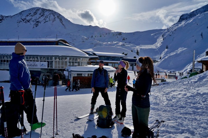 Group of people in winter gear standing on snowy ski slope with mountains and ski lift in background.