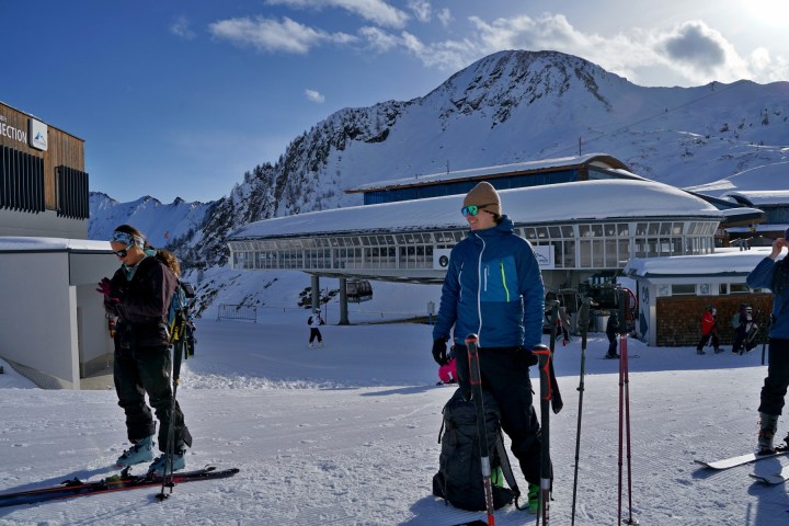 Skiers near a snow-covered mountain and ski resort building with ski poles and backpacks.
