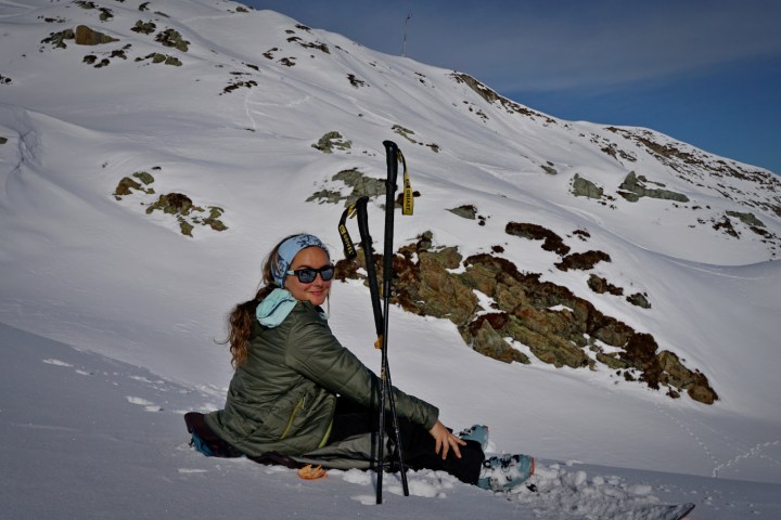 Person sitting on snow with ski poles, wearing sunglasses and a headband, snowy mountain background.