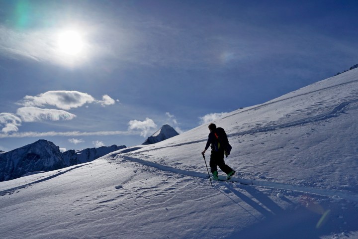 Silhouette of a person with poles hiking on a snowy mountain under a bright sun and blue sky.
