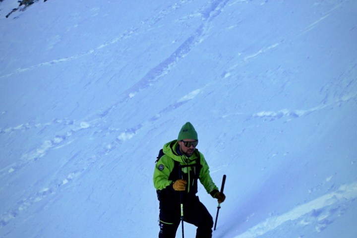 Person in green jacket climbing snowy mountain with poles.