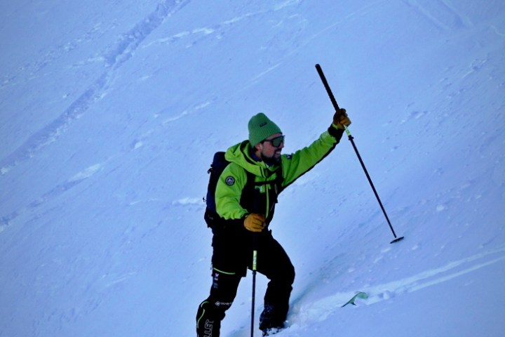 Person in green jacket and hat climbing a snowy slope with ski poles.