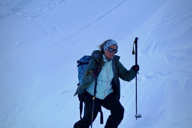 Person skiing downhill on a snowy slope with poles and backpack, wearing goggles and a headband.