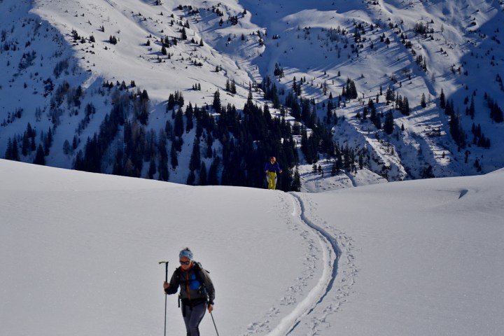 A person skiing uphill on a snowy mountain with another skier in the background under clear blue sky.