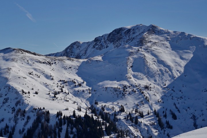 Person skiing up a snow-covered mountain with clear blue sky and airplane contrails.