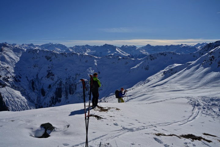 Two skiers on a snowy mountain peak with ski poles, surrounded by alpine scenery.