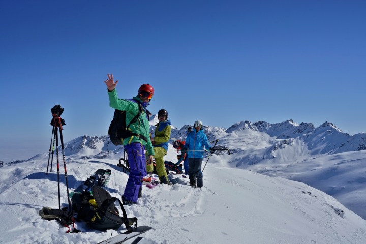 Skiers on snowy mountain peak with gear, clear blue sky background.