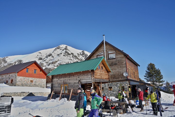 People in winter gear outside a log cabin with snowy mountains in the background.