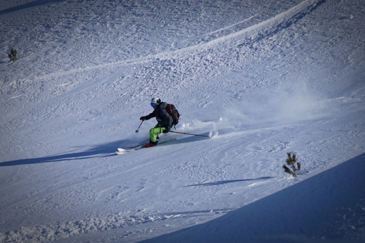 Skier in green pants glides down a snowy slope with poles, snow spraying behind.