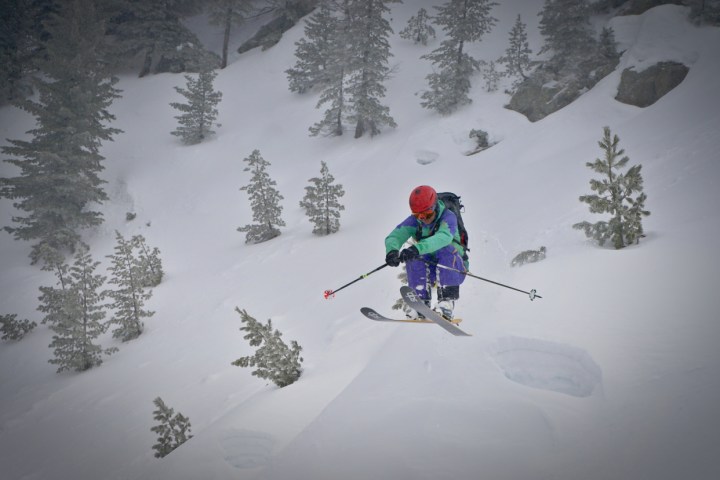 Skier in colorful gear jumps off a snowy slope amid pine trees.