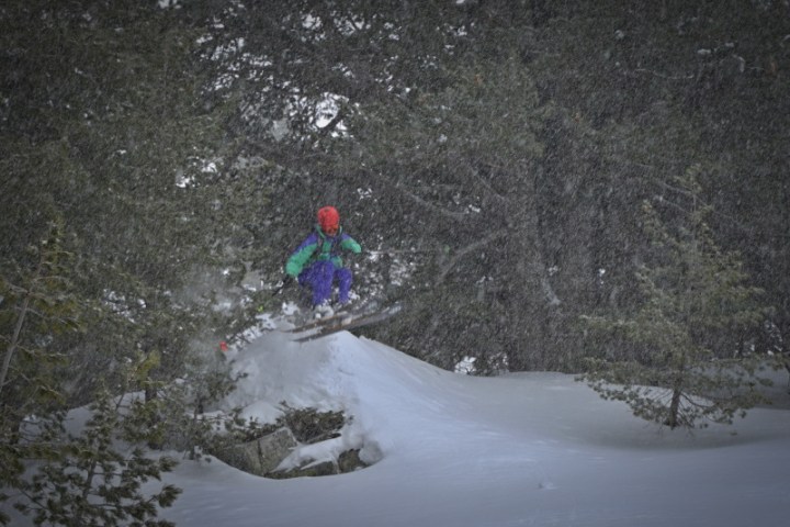 Skier in colorful gear jumps off a snowy hill in a forest during snowfall.