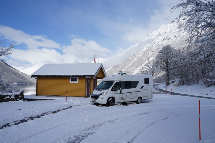 Campervan parked in snow by a yellow cabin with snowy mountains and trees in the background.