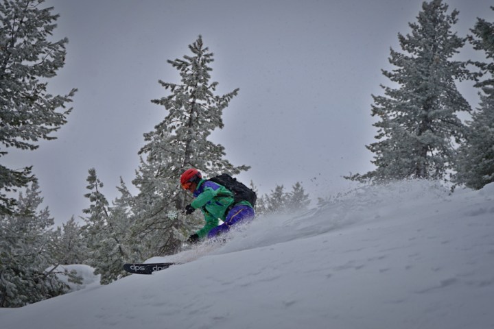 Person in colorful gear skiing downhill through powdery snow, surrounded by snow-covered trees.