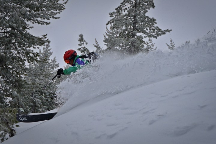 Skier in colorful gear descending a snowy slope with trees in the background.