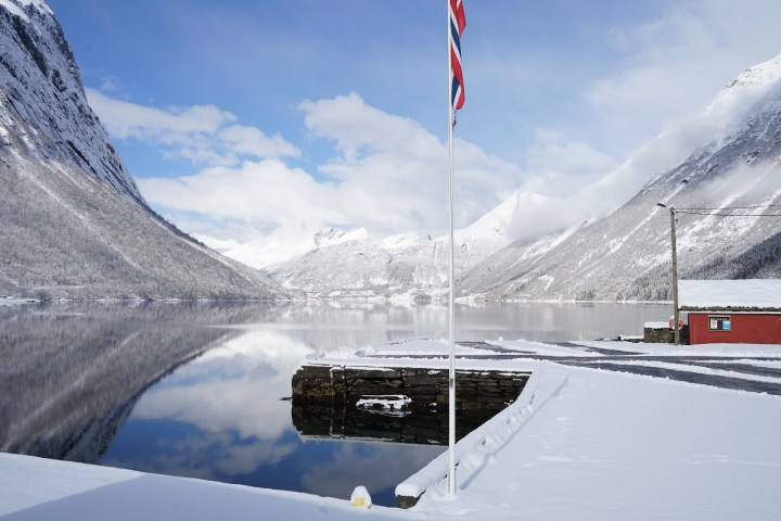 Snowy fjord landscape with Norwegian flag, red building, and mountains reflected in water.