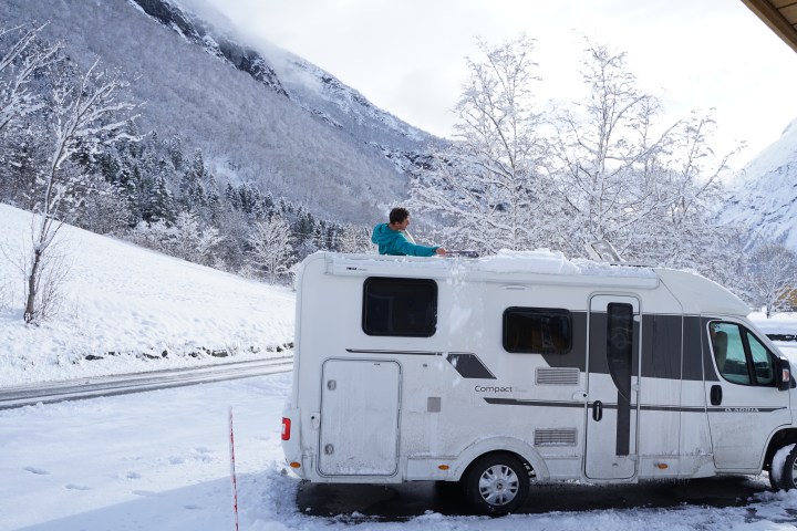 Person clearing snow off RV roof in snowy mountain landscape.