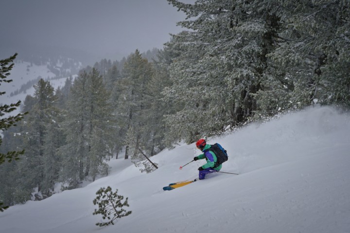 Skier in colorful gear descends a snowy slope surrounded by pine trees.
