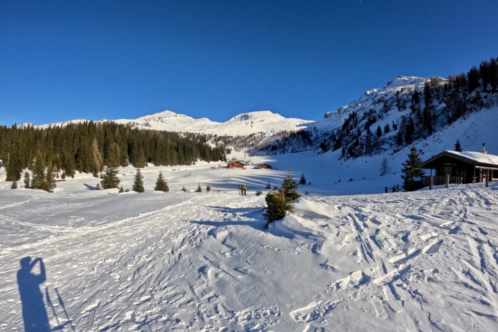 Snowy landscape with trees, mountains, and a cabin under a clear blue sky.