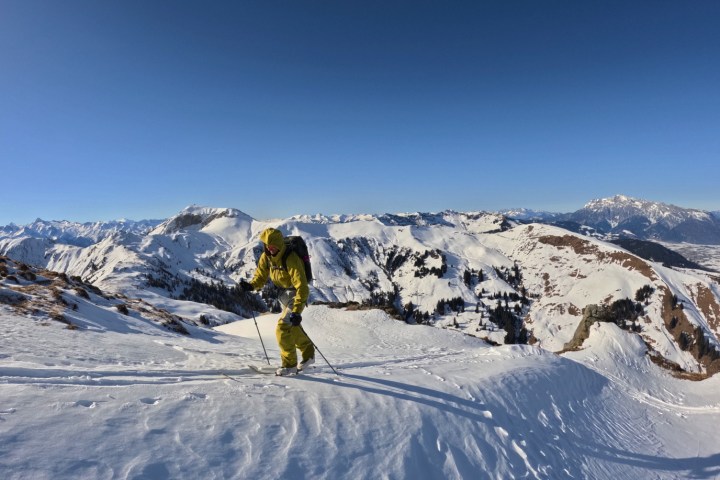 Person in yellow suit skiing uphill on a snowy mountain with a clear blue sky and distant peaks.