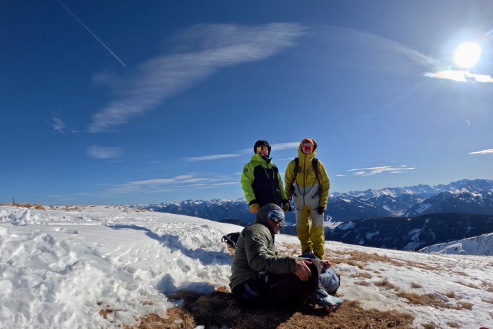 Three people in ski gear on a snowy mountain with a clear blue sky and sun.