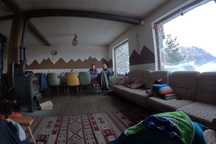 Cozy room with mountain decor, two people at table, couches, stove, large windows showing snowy landscape.