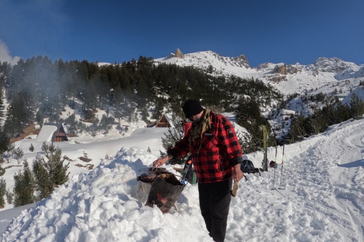 Person in red plaid jacket tending to a snowy fire pit, with mountain and trees in the background.