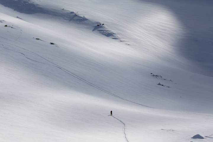 Lone skier on vast snowy slope with ski tracks and rocky patches.
