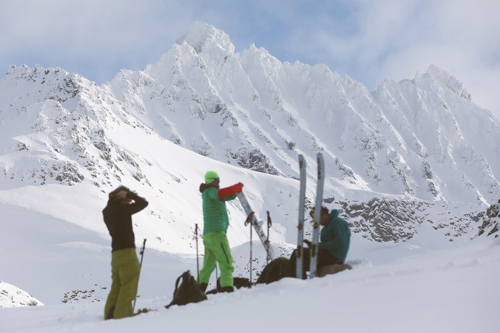 Skiers preparing on a snowy mountain slope with large craggy peaks in the background.