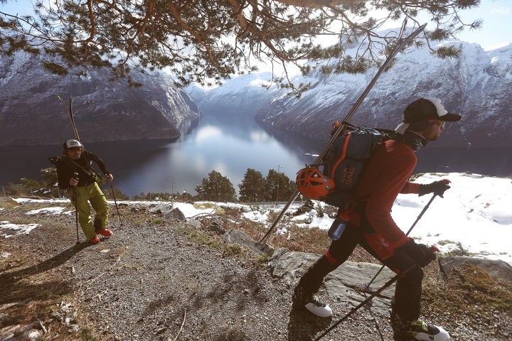 Two hikers with ski gear walk on a snowy trail overlooking a fjord and mountains.