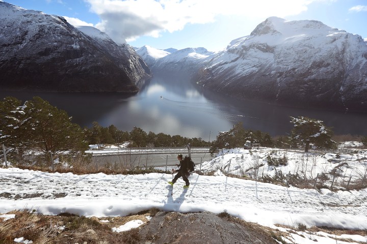 Person hiking up a snowy path with a lake and snow-covered mountains in the background.