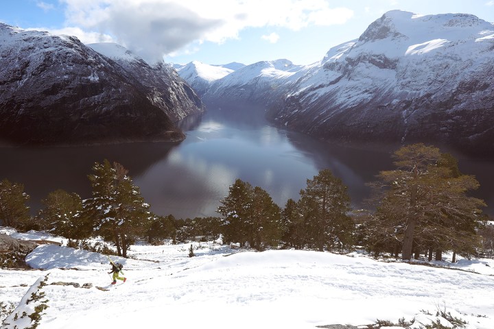 Person hiking up snowy hill with fjord and snow-covered mountains in the background.