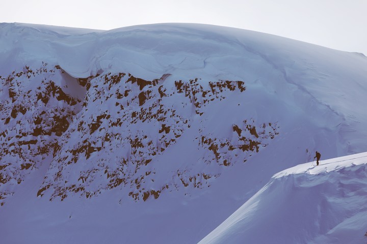 Person skiing on a snowy mountain ridge with rocky slopes in the background.