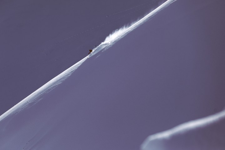Skier descending a steep snowy slope, leaving a trail of snow dust in a vast, snow-covered landscape.