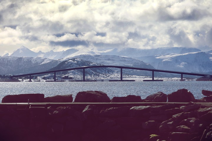 Snow-capped mountains with a bridge over calm water under cloudy skies.