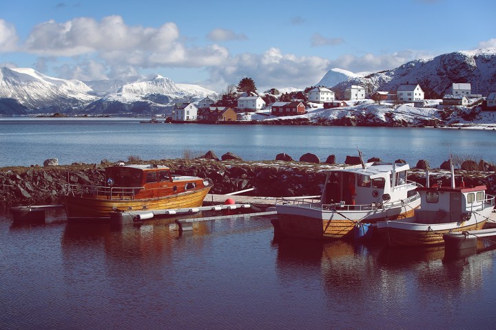 Boats docked in a snowy harbor with mountains and village houses in the background.