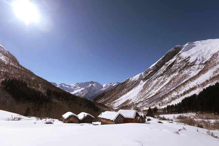 Snowy mountain landscape with wooden cabins and bright sun in clear blue sky.
