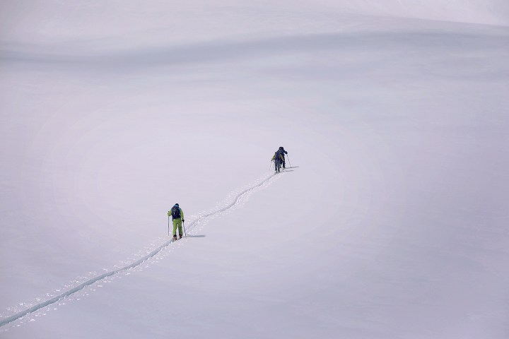 Two skiers traverse a vast snowy landscape, leaving a trail behind them.