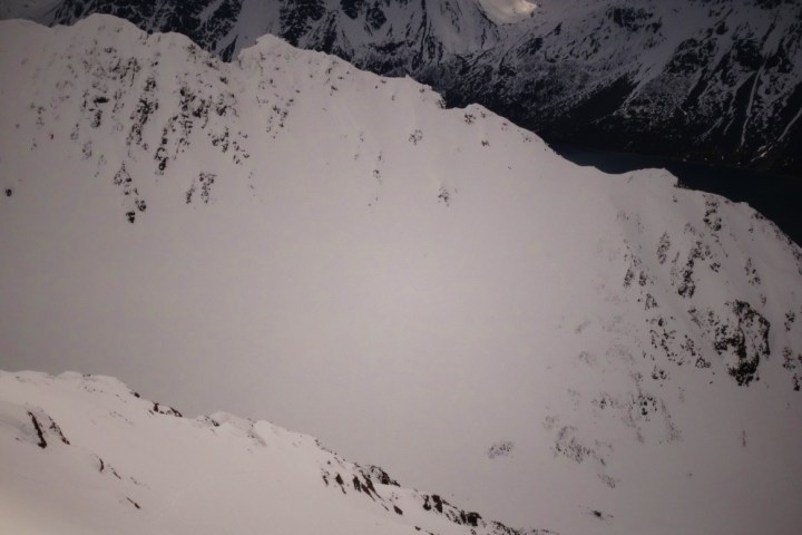 Two people hike on snowy mountain with skis, large jagged peaks in background.