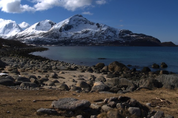 Snowy mountains by a rocky shoreline with calm water under a blue sky.