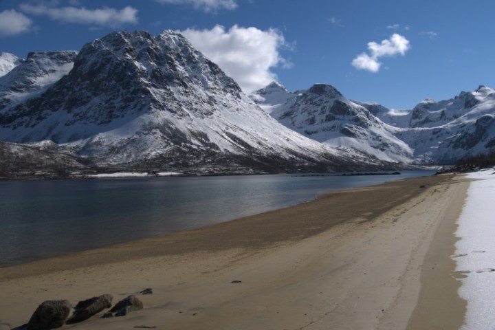 Snow-capped mountains by a calm sea, with a sandy beach in the foreground under a clear blue sky.