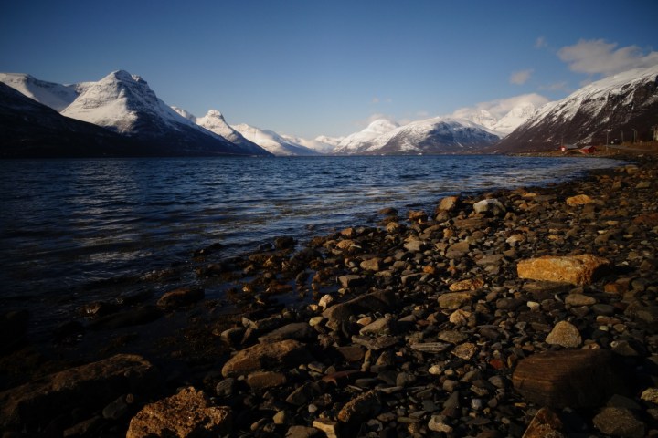 Rocky shoreline with snow-capped mountains and blue sky in the background.