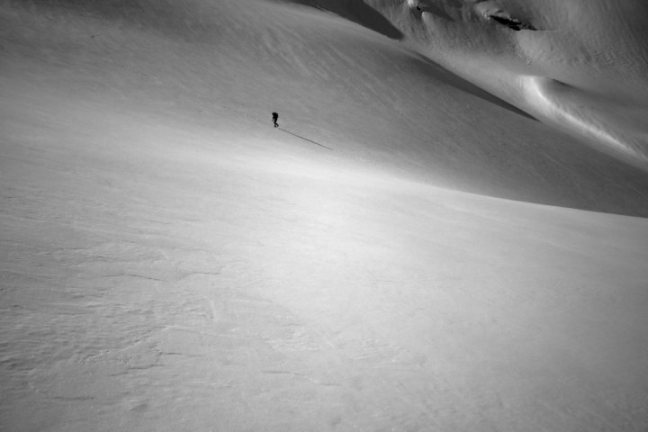 Lone skier traversing a vast, smooth snow-covered mountain slope under a clear sky.