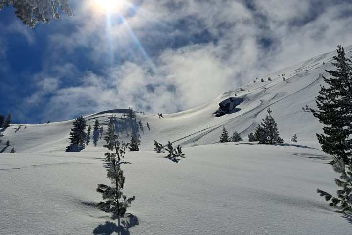 Snowy mountain landscape with sun, trees, and ski tracks under a partly cloudy sky.