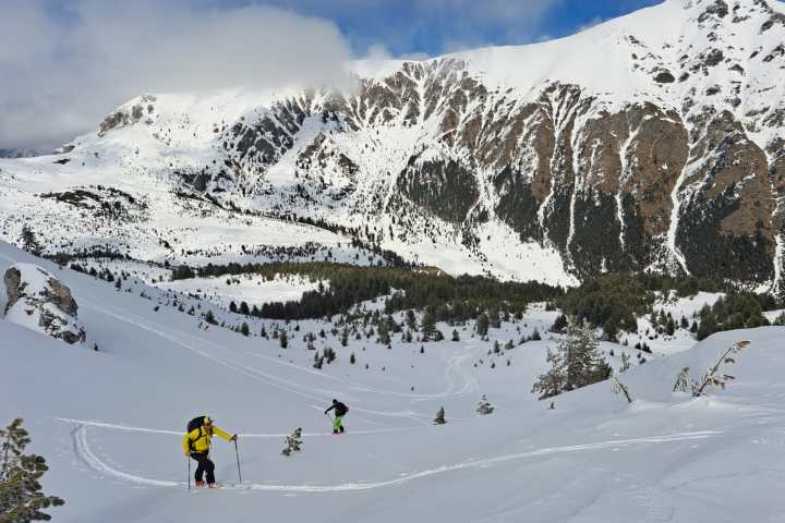 Ski tourers ascending a snowy mountain slope with rocky peaks in the background under a cloudy sky.