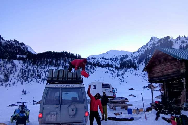 People loading a van at a snowy mountain camp at dusk with tents and a small cabin nearby.