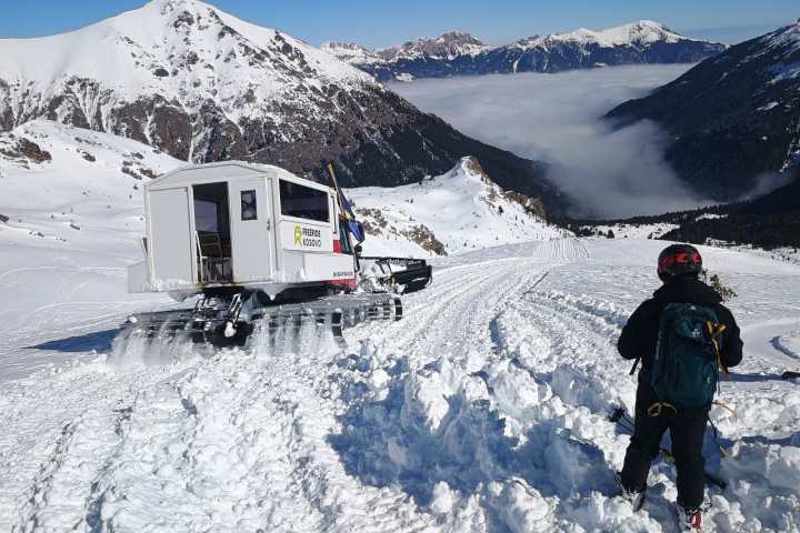 Person with gear looks at snowcat on snowy mountain trail under clear blue sky.