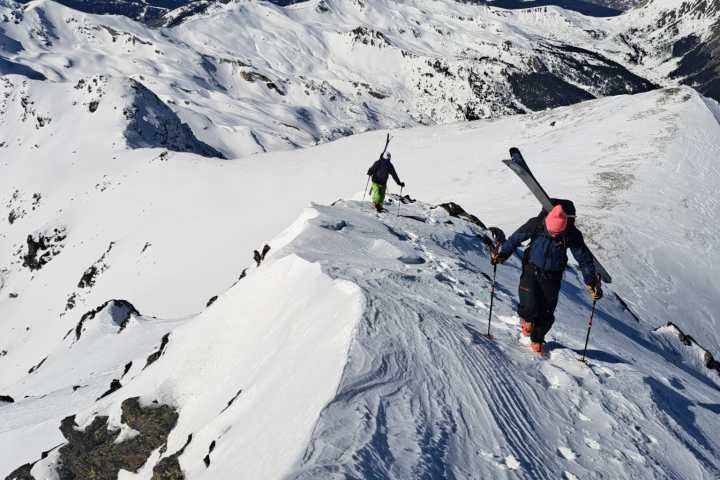 Two skiers with poles ascend a snowy mountain ridge under a clear blue sky.