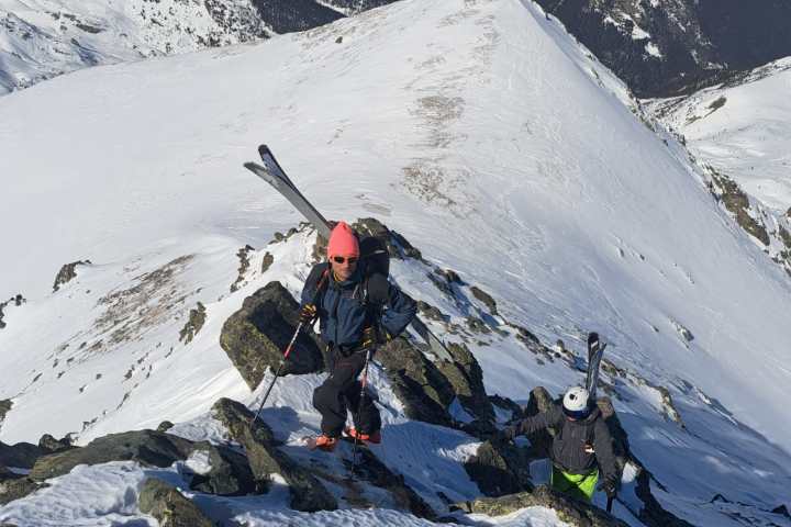 Two skiers hiking up a snowy mountain ridge with skis on their backpacks.