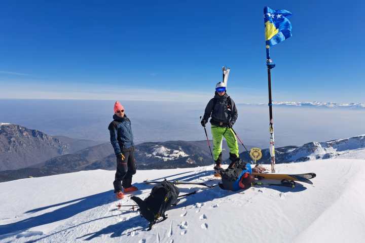 Two skiers on a snowy mountain summit with a flag, bright blue sky in the background.